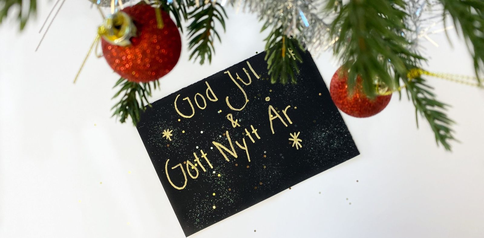 Närbild av julgran med röda glittriga kulor och ett svart kort med texten God Jul & Gott Nytt År